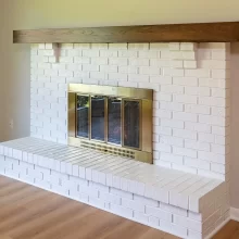 White painted brick fireplace with raised hearth, brass fireplace doors, wood mantel, and light wood flooring