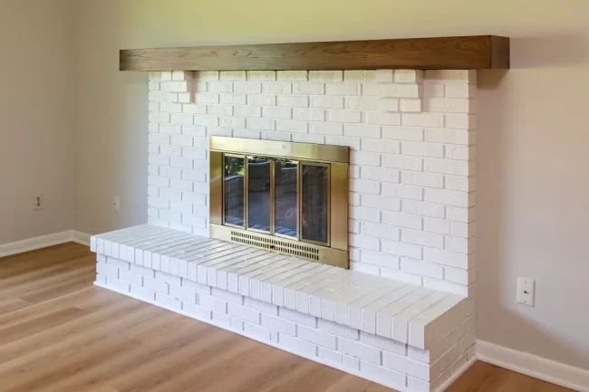 White Brick Fireplace with Wood Mantel | Pennings & Sons White painted brick fireplace with raised hearth, brass fireplace doors, wood mantel, and light wood flooring