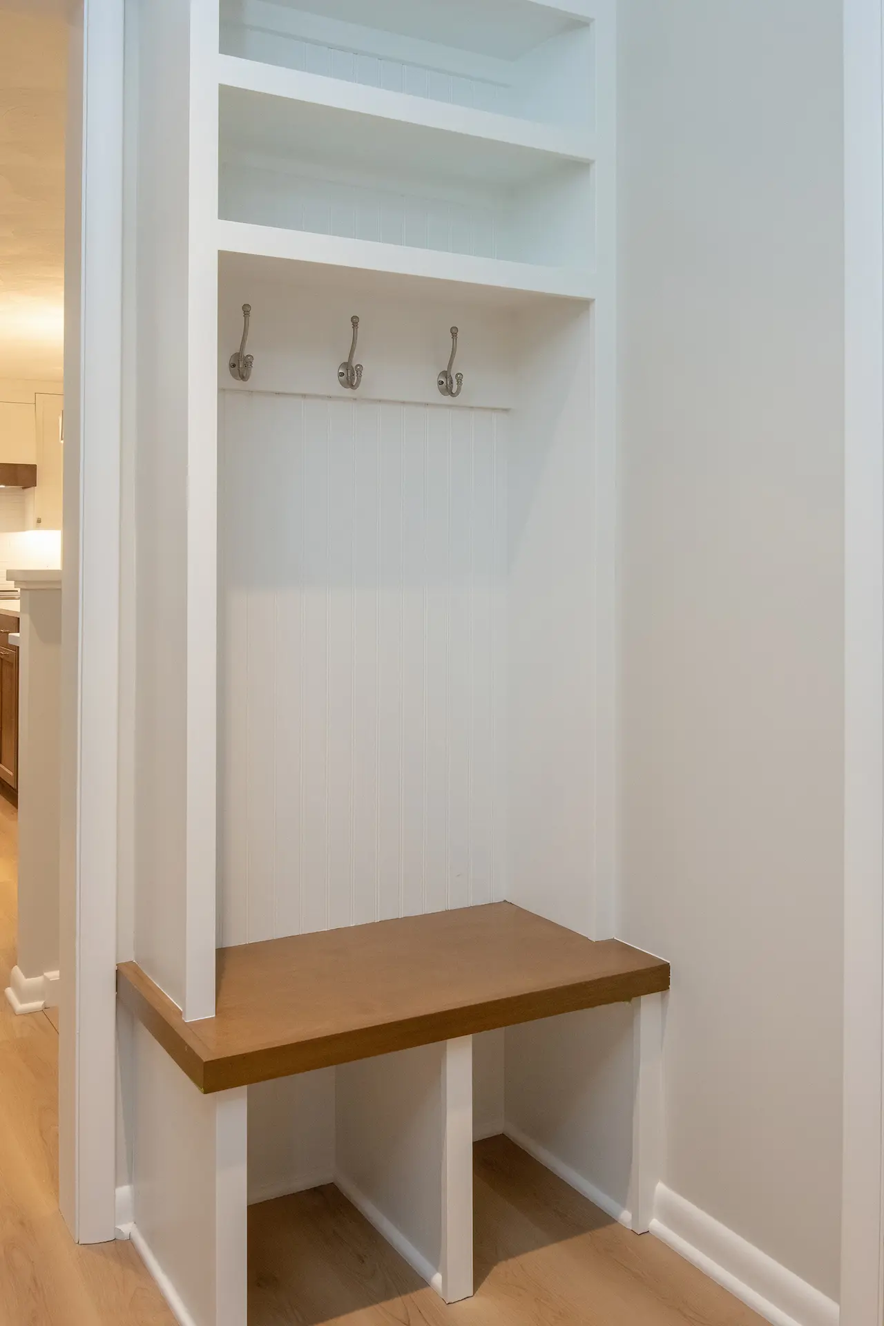 Built-in mudroom bench with wood seat, white beadboard backing, coat hooks, open cubbies, and upper storage shelves.