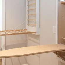 Wall-mounted fold-down drying rack closed against a beige wall beside a white cabinet and light wood flooring.