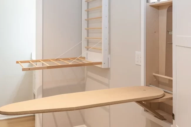 Wall-Mounted Fold-Down Drying Rack | Pennings & Sons Wall-mounted fold-down drying rack closed against a beige wall beside a white cabinet and light wood flooring.