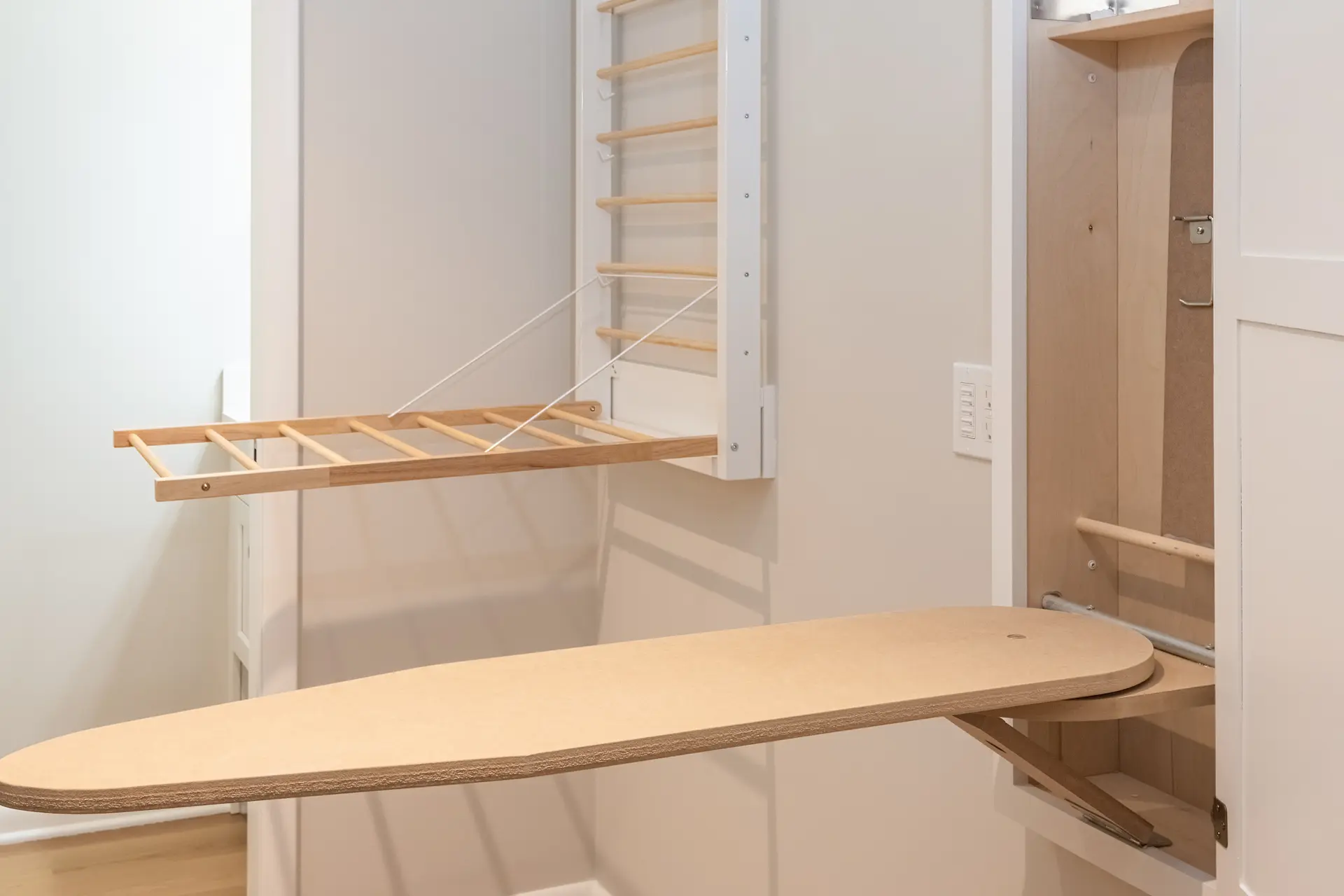 Wall-mounted fold-down drying rack closed against a beige wall beside a white cabinet and light wood flooring.