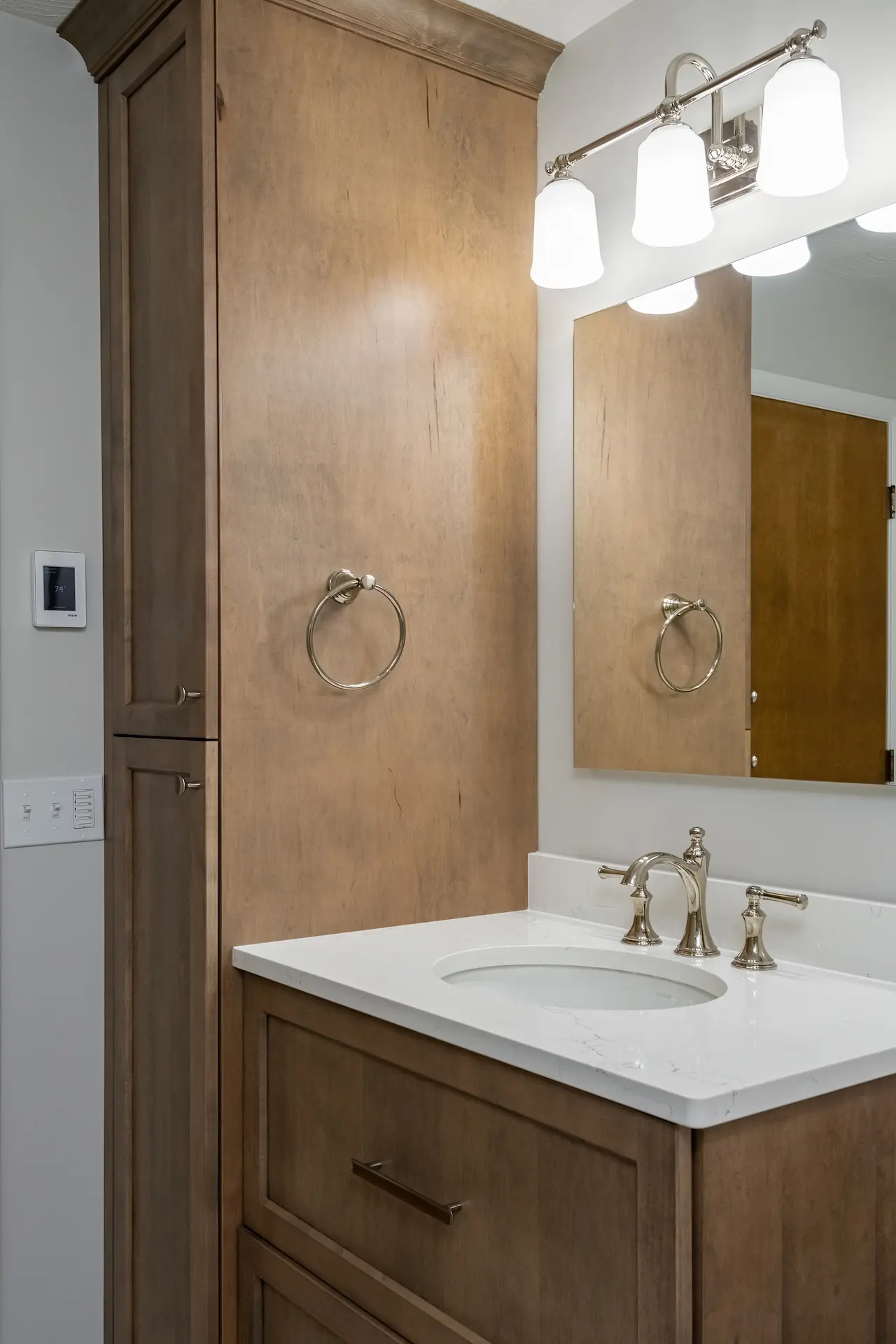 Wood bathroom vanity with tall linen cabinet, white countertop, polished nickel faucet, mirror, and three-light vanity fixture