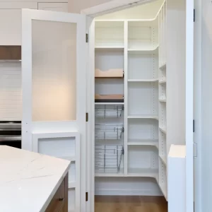 Open kitchen pantry with white shelving, pull-out wire baskets, frosted glass door, and nearby quartz kitchen island