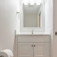 Small bathroom vanity with white cabinet, white countertop, mirror, three-light fixture, and open lower storage shelf.