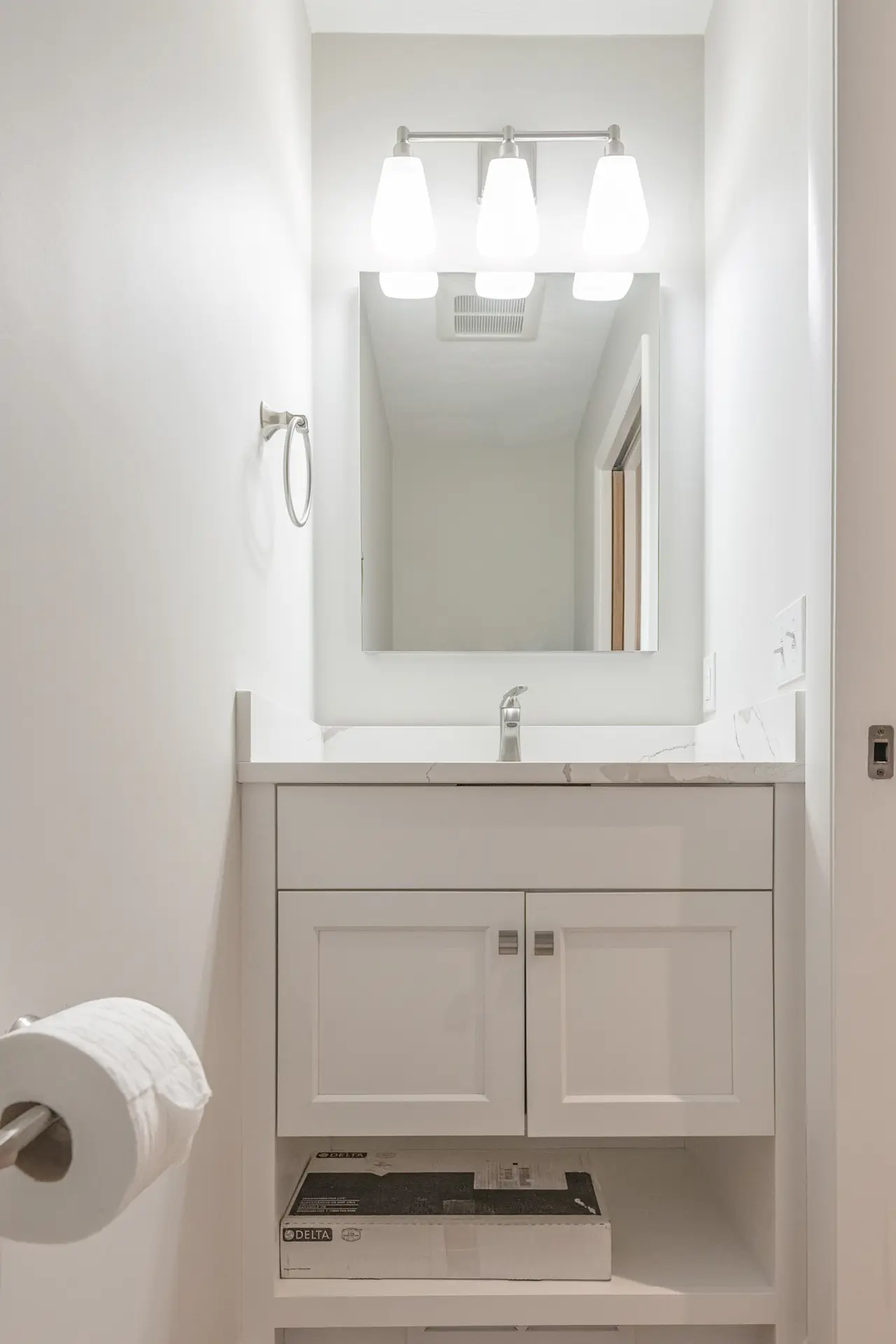Small bathroom vanity with white cabinet, white countertop, mirror, three-light fixture, and open lower storage shelf.