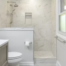 Bathroom with white marble-look tile walk-in shower, half wall, toilet, light gray floor tile, and window