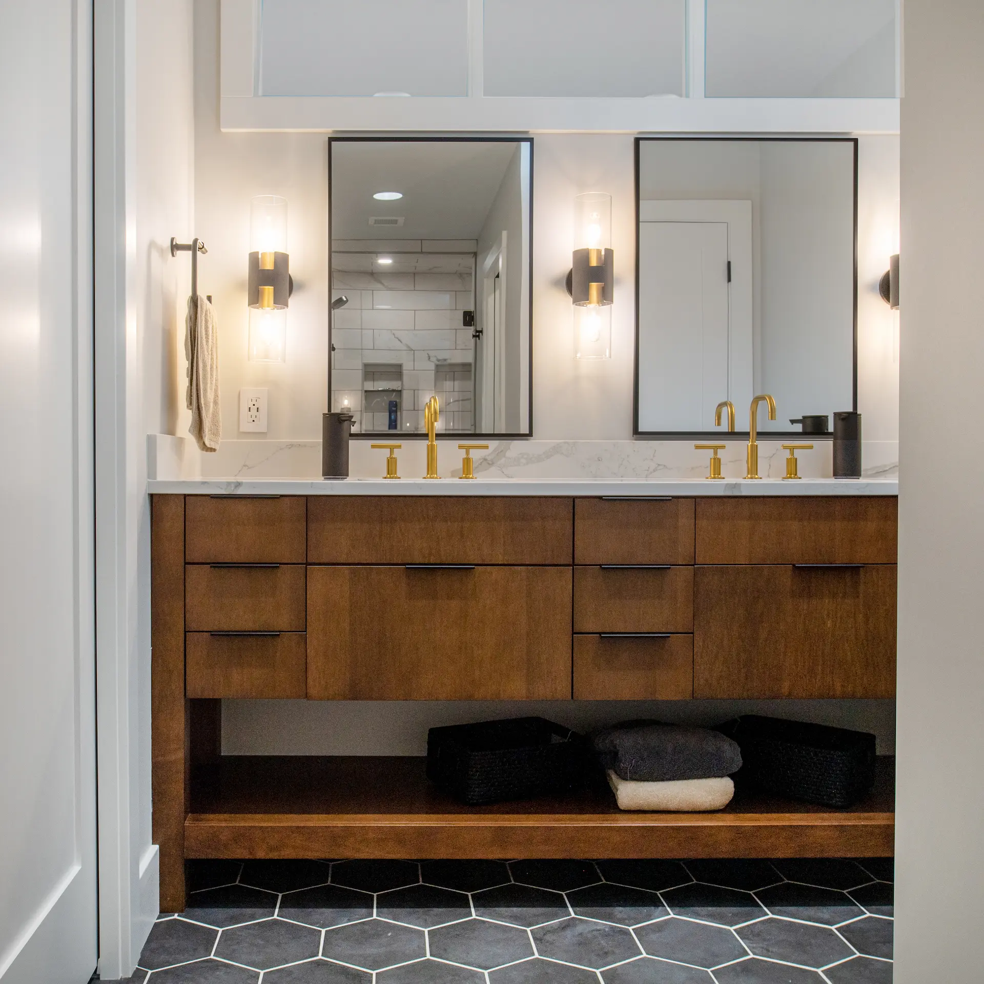 Bathroom with floating wood double vanity, gold faucets, wall sconces, black hex tile floor, and custom mirrors