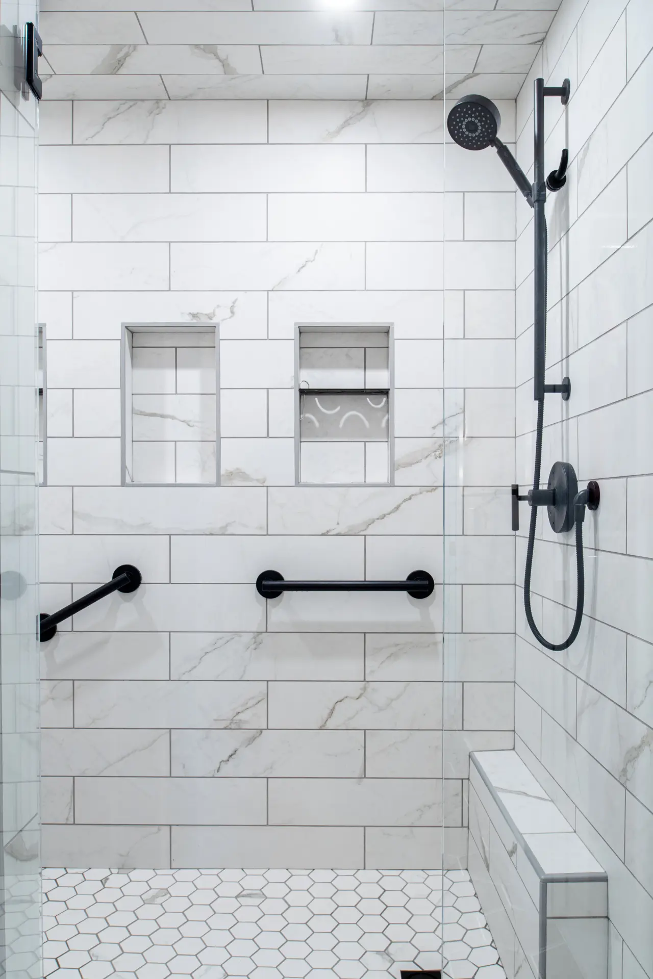 Walk-in shower with marble-look tile, glass enclosure, black fixtures, grab bars, and built-in niches