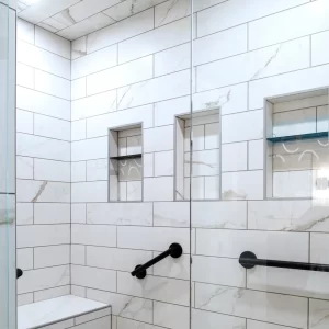 Close-up of marble-look shower tile with recessed niches, glass enclosure, black fixtures, and shower bench