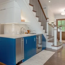Blue wet bar cabinetry with white counters built beneath a wood staircase
