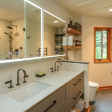 Bathroom with double vanity, lighted mirrors, open shelving, woven baskets, tile floor, and glass shower reflection