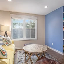 Loft sitting area with yellow sofa, blue built-in game shelves, large window, wood flooring, and round coffee table