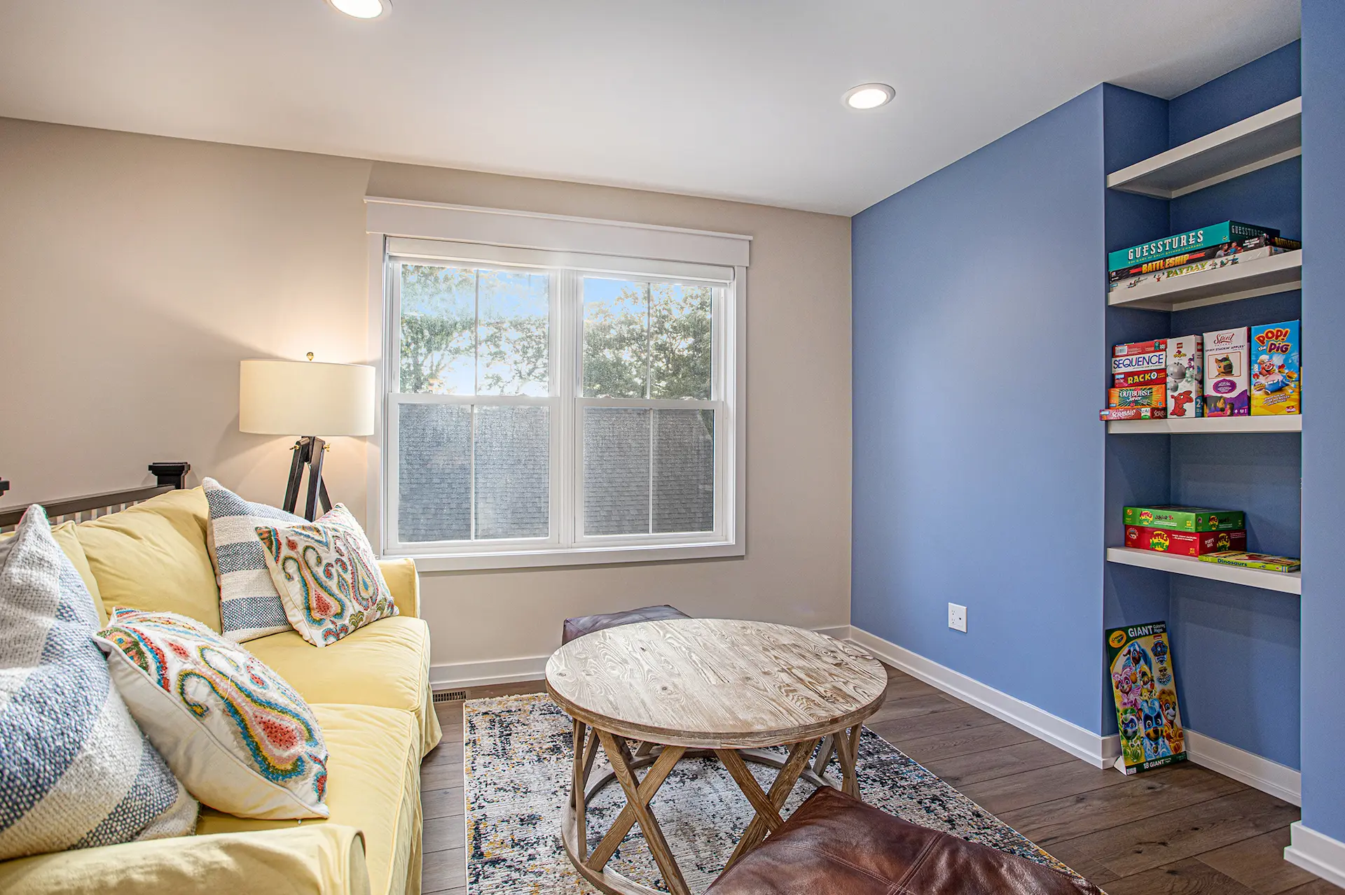 Loft sitting area with yellow sofa, blue built-in game shelves, large window, wood flooring, and round coffee table