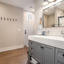 Bathroom with long gray double vanity, black fixtures, wall mirror, entry hooks, and shower beyond the doorway