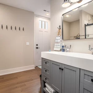 Bathroom with long gray double vanity, black fixtures, wall mirror, entry hooks, and shower beyond the doorway
