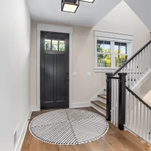 Interior entryway with black front door, wood flooring, round patterned rug, window, and staircase with black and white railing