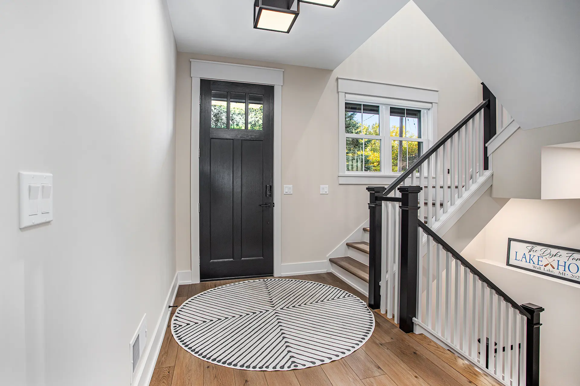 Interior entryway with black front door, wood flooring, round patterned rug, window, and staircase with black and white railing
