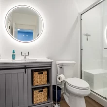 Guest bathroom with gray vanity, round lighted mirror, toilet, and glass shower enclosure