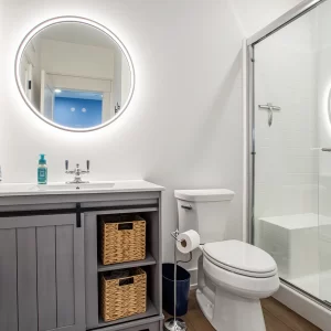 Guest bathroom with gray vanity, round lighted mirror, toilet, and glass shower enclosure