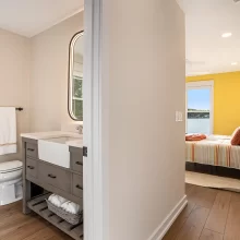 Guest bathroom with gray vanity, farmhouse sink, illuminated mirror, black hardware, toilet, window, and glass shower enclosure