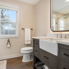 Guest bathroom with gray vanity, farmhouse sink, illuminated mirror, black hardware, toilet, window, and glass shower enclosure