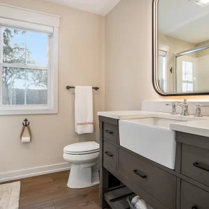 Guest bathroom with gray vanity, farmhouse sink, illuminated mirror, black hardware, toilet, window, and glass shower enclosure