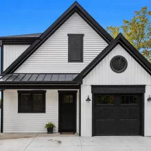 Front exterior of modern lakefront home with white siding, black trim, black garage door, round gable window, concrete driveway, and lake view to the side