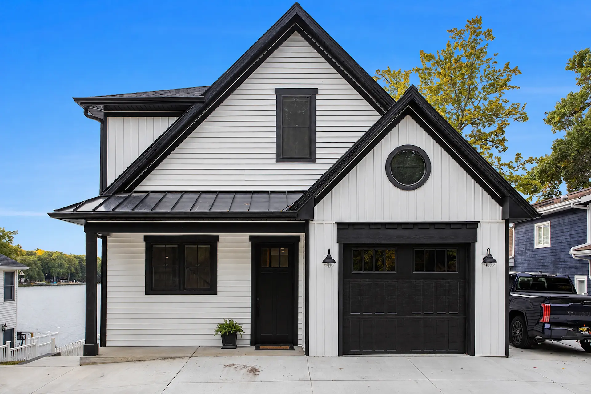 Front exterior of modern lakefront home with white siding, black trim, black garage door, round gable window, concrete driveway, and lake view to the side