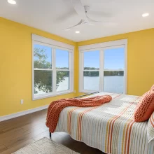 Bright bedroom with yellow accent walls, lake views through corner windows, striped bedding, wood flooring, and white ceiling fan