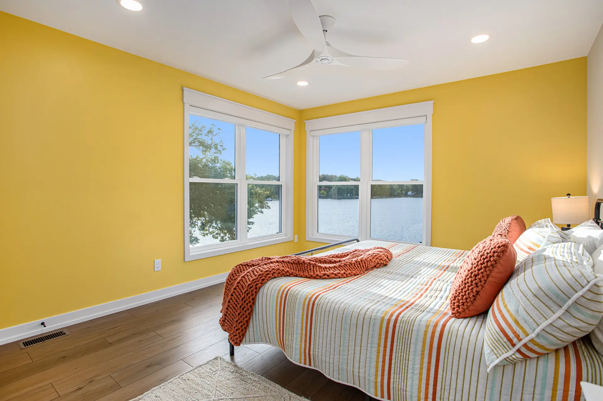 Bright bedroom with yellow accent walls, lake views through corner windows, striped bedding, wood flooring, and white ceiling fan