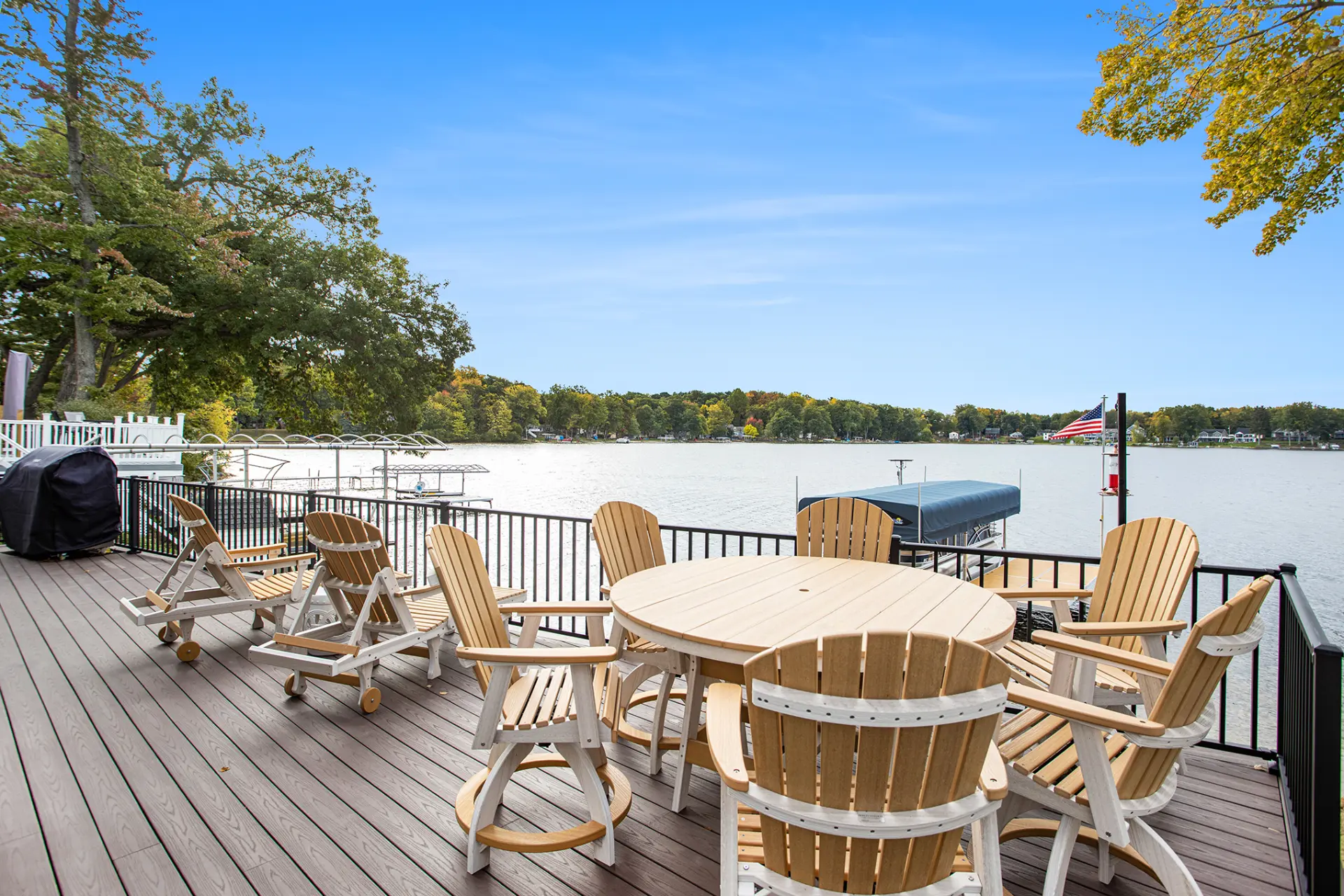 Lakefront deck with outdoor dining table, Adirondack chairs, black railing, lake view, dock, and blue sky