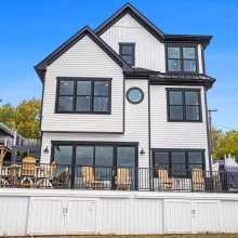 Back exterior of lakefront home with large deck, outdoor seating, black railings, white siding, and lake-facing windows
