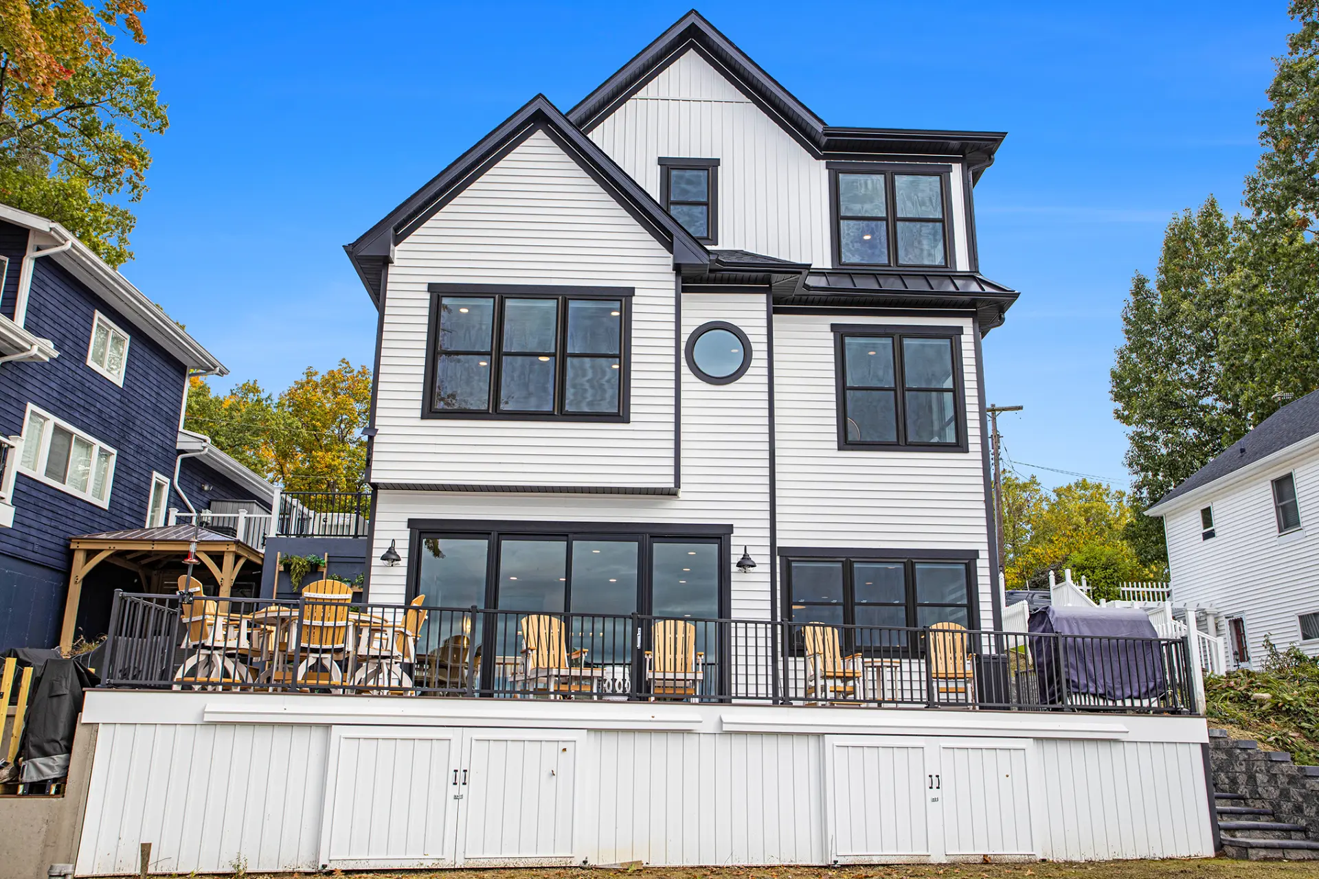 Back exterior of lakefront home with large deck, outdoor seating, black railings, white siding, and lake-facing windows