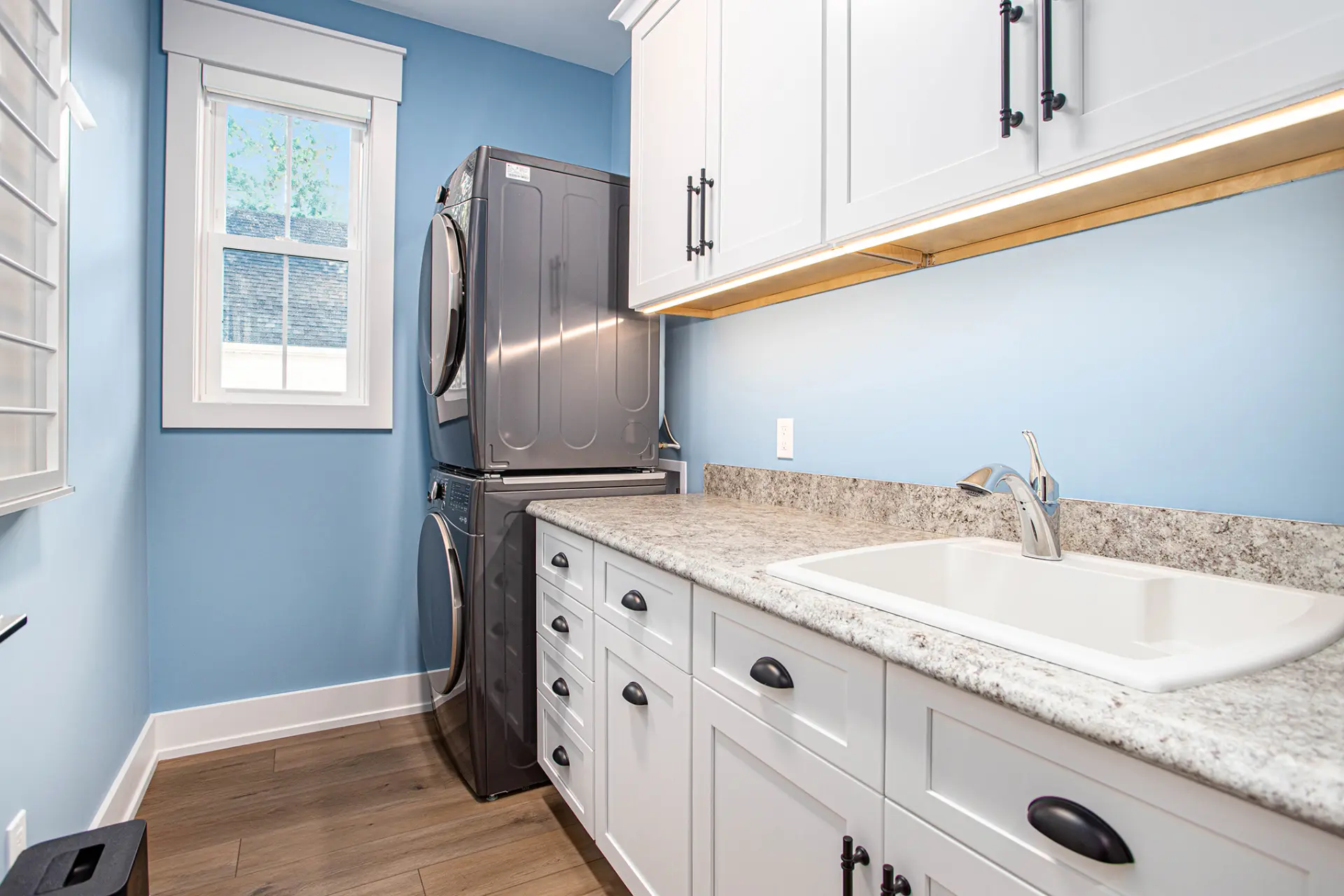Laundry room with stacked washer and dryer, white cabinets, granite countertop, utility sink, and blue walls