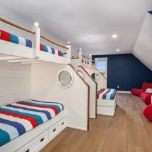 Nautical bunk room with built-in loft beds, rope railings, striped bedding, red lounge chairs, blue accent wall, and wood flooring