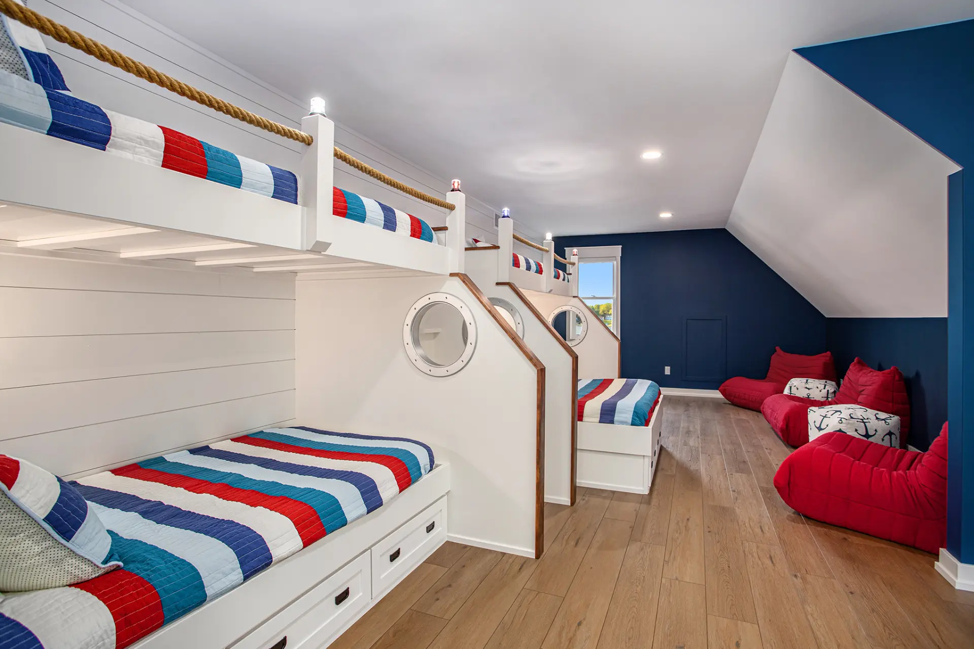 Nautical bunk room with built-in loft beds, rope railings, striped bedding, red lounge chairs, blue accent wall, and wood flooring