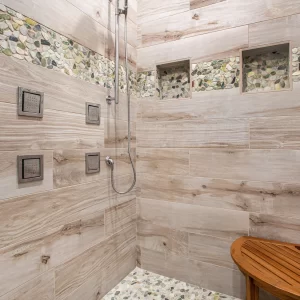 Walk-in shower with wood-look wall tile, pebble accent band, built-in niches, body sprays, and teak bench