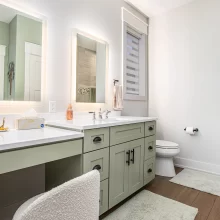 Bathroom with sage green double vanity, illuminated mirrors, white countertops, toilet, and wood-look tile shower