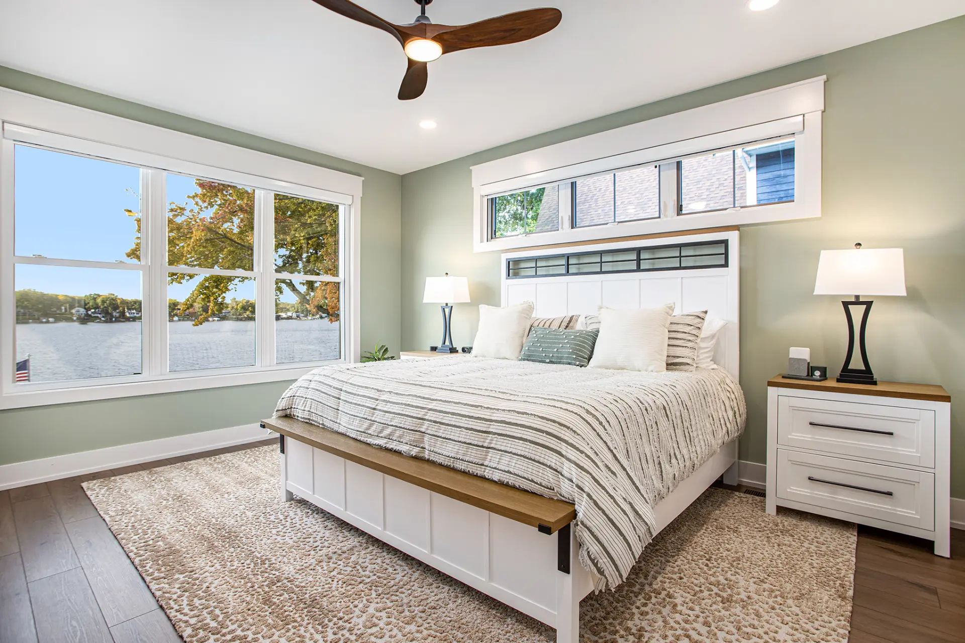 Primary bedroom with green accent wall, white bed frame, transom windows, lake views, and ceiling fan
