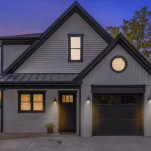 Modern lakefront home exterior at twilight with white siding, black trim, black garage door, warm window lighting, and lake view beside the house