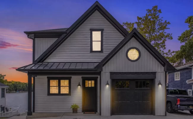 Modern Lakefront Home Twilight Exterior | Pennings & Sons Modern lakefront home exterior at twilight with white siding, black trim, black garage door, warm window lighting, and lake view beside the house