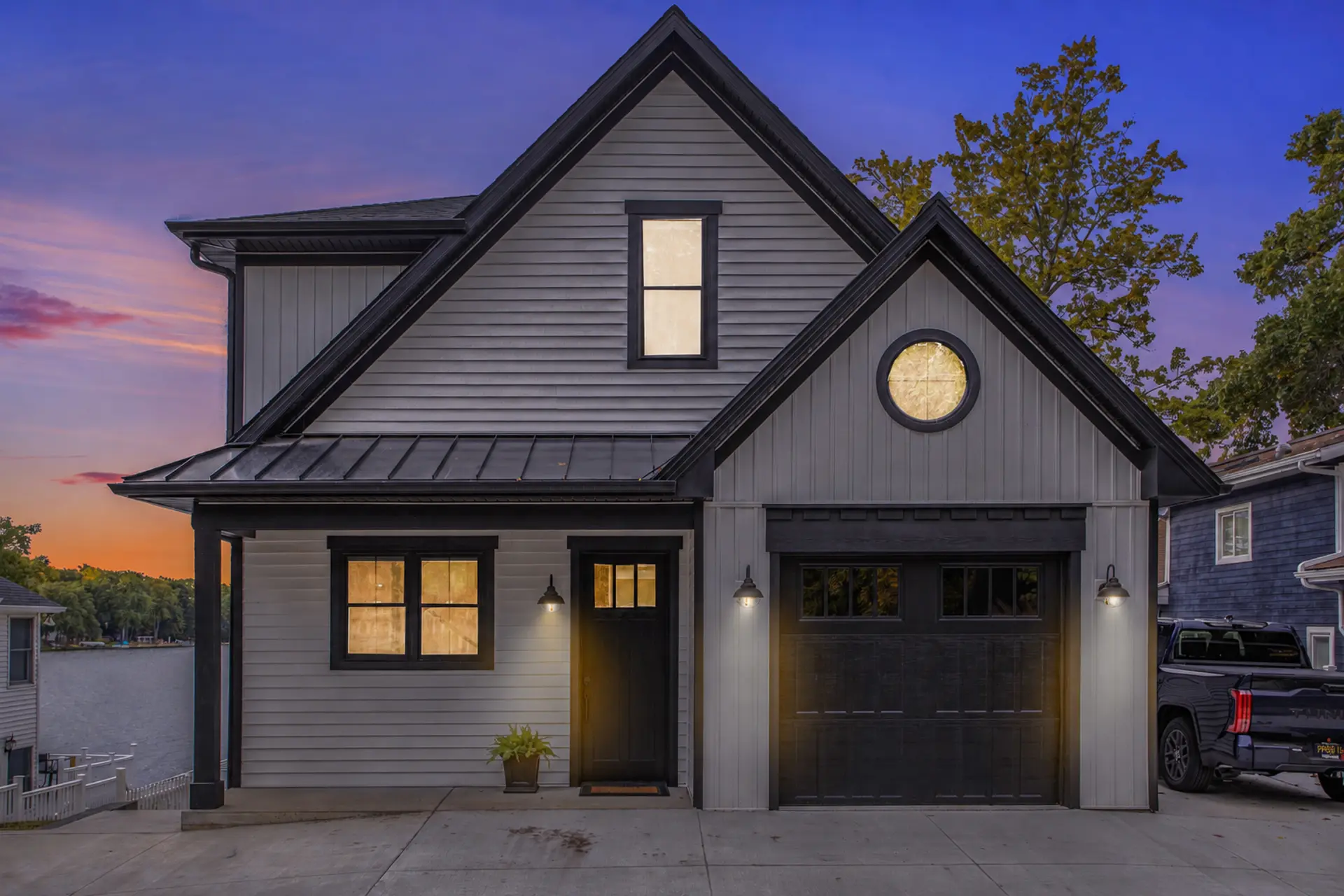Modern lakefront home exterior at twilight with white siding, black trim, black garage door, warm window lighting, and lake view beside the house
