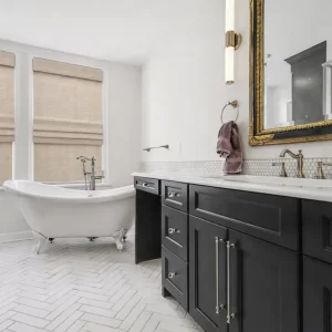 Primary bathroom with black double vanity, ornate gold mirror, white clawfoot tub, glass shower, and white herringbone tile floor in a downtown Kalamazoo condominium
