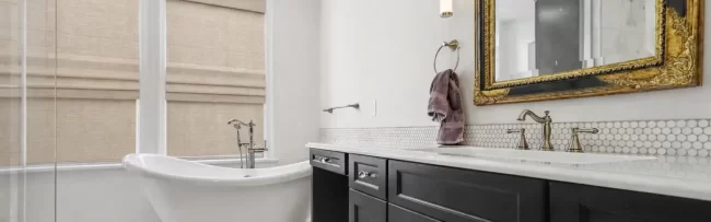 Primary bathroom with black double vanity, ornate gold mirror, white clawfoot tub, glass shower, and white herringbone tile floor in a downtown Kalamazoo condominium