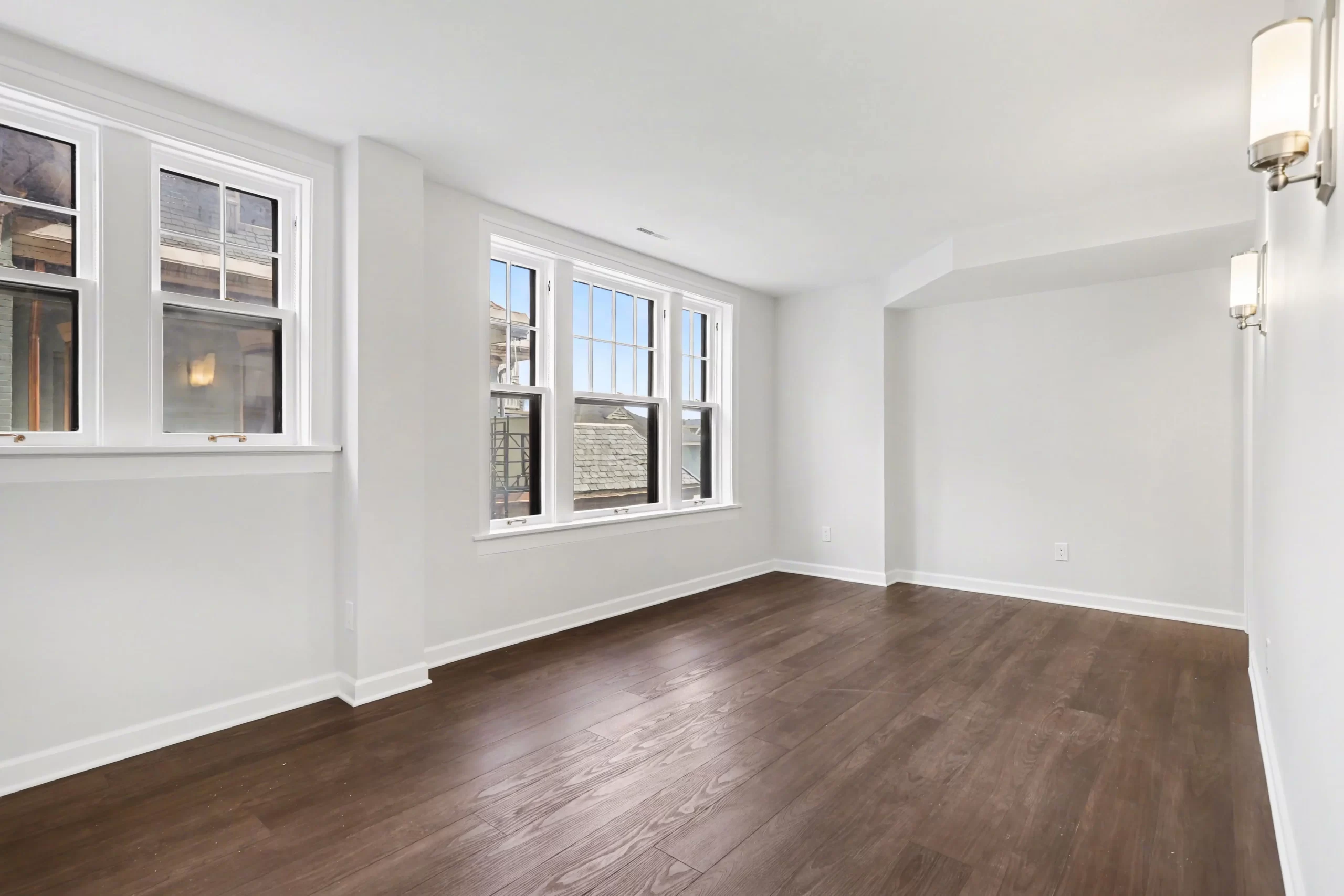Unfurnished room with dark hardwood flooring, white trim, wall sconces, and large windows in a downtown Kalamazoo condominium