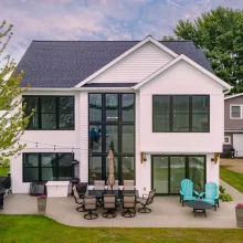 Rear exterior of lakefront home with large black-framed windows, outdoor dining area, patio seating, and lake view