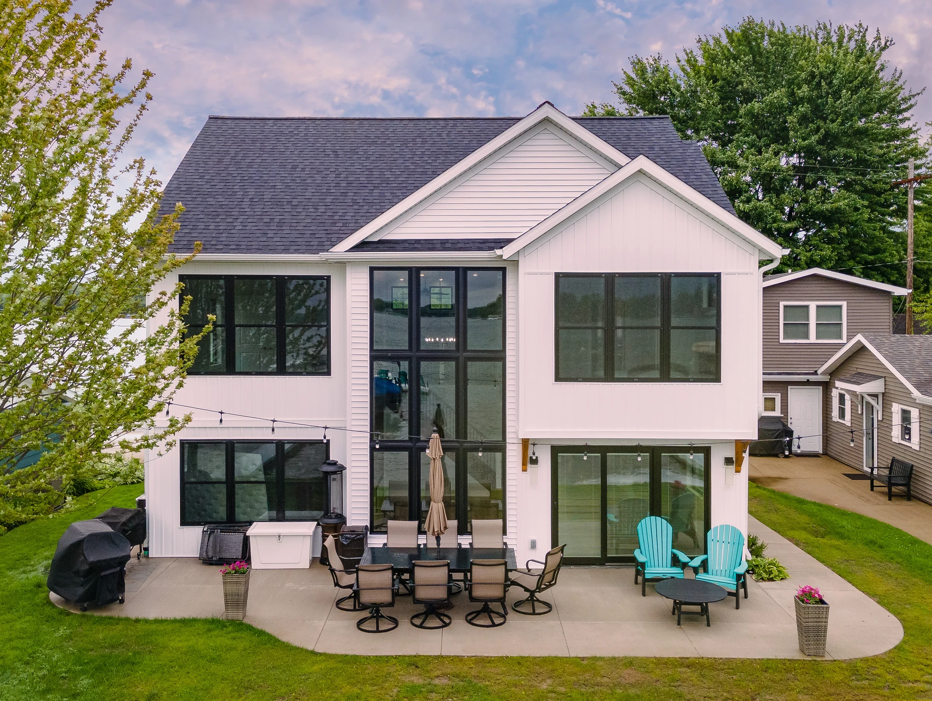 Rear exterior of lakefront home with large black-framed windows, outdoor dining area, patio seating, and lake view
