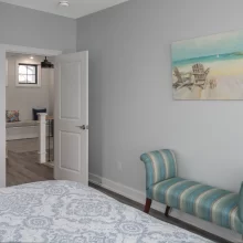 Bedroom with gray walls, patterned bedding, striped accent bench, beach artwork, and doorway to adjoining sitting area
