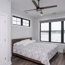 Bedroom with black-framed lake view windows, wood bed frame, patterned bedding, ceiling fan, and dark wood-look flooring